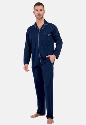 Man met kort haar die marineblauw, langärmelig pyjama met witte biezen draagt, staat blootsvoets met één hand in de zak.