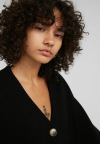 Schwarze gerippte Strickjacke mit tiefem V-Ausschnitt, ausgestattet mit einem großen Knopfdetail, getragen präsentiert, wobei die Textur und die lässige Passform hervorgehoben werden.