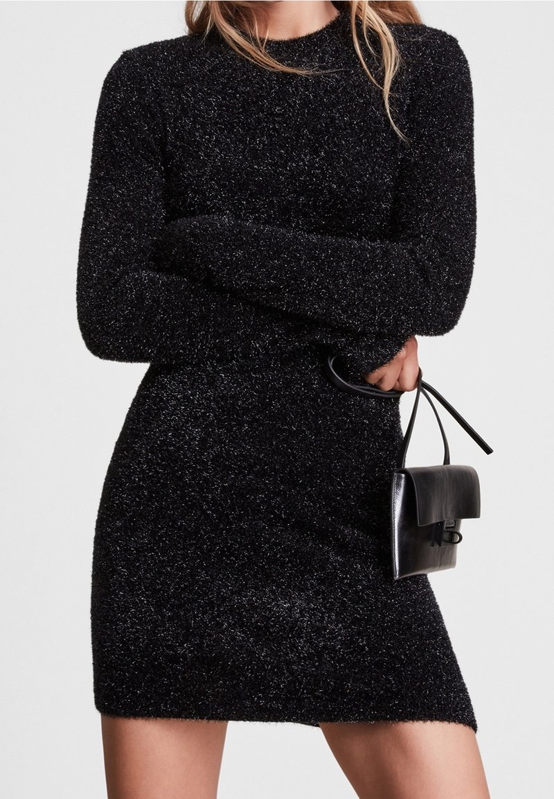 Femme portant une robe noire en peluche à manches longues, tenant un petit sac à main en cuir noir par sa poignée sur un fond uni.