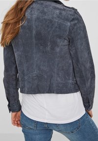 Grå mockajacka med en kortad design, utrustad med sömnadsdetaljer, framfickor med lock och långa ärmar. Kombinerad med en vit skjorta under.