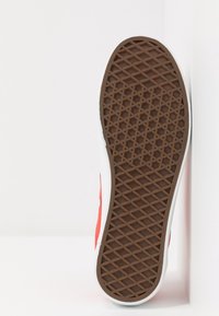 Semelle de chaussure présentant une partie inférieure en caoutchouc brun texturé avec un motif en losanges, bordée d'un bord synthétique blanc et d'une tige en toile rouge.