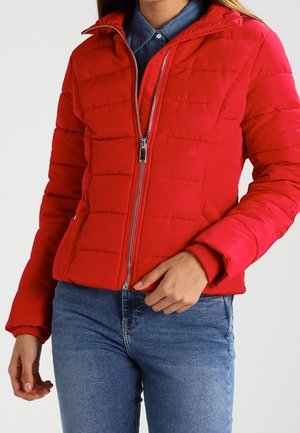 Femme portant une veste matelassée rouge avec une fermeture éclair devant, sur une chemise en jean bleue et un jean bleu, main près de la fermeture éclair de la veste.
