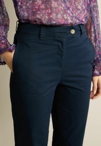 Pantaloni blu navy in un tessuto liscio con tasche laterali, chiusura con bottone e sottili particolari di cucitura, abbinati a un top floreale e trasparente.