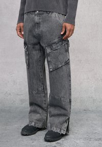 Pantalones cargo en denim negro deslavado con múltiples bolsillos, bordes desgastados y un corte holgado. Presenta el texto bordado "Utopia" en la parte frontal.