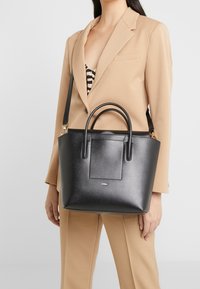 Svart läderhandväska med dubbla handtag upptill, en avtagbar axelrem och en platt framficka. Slät textur och strukturerad form.