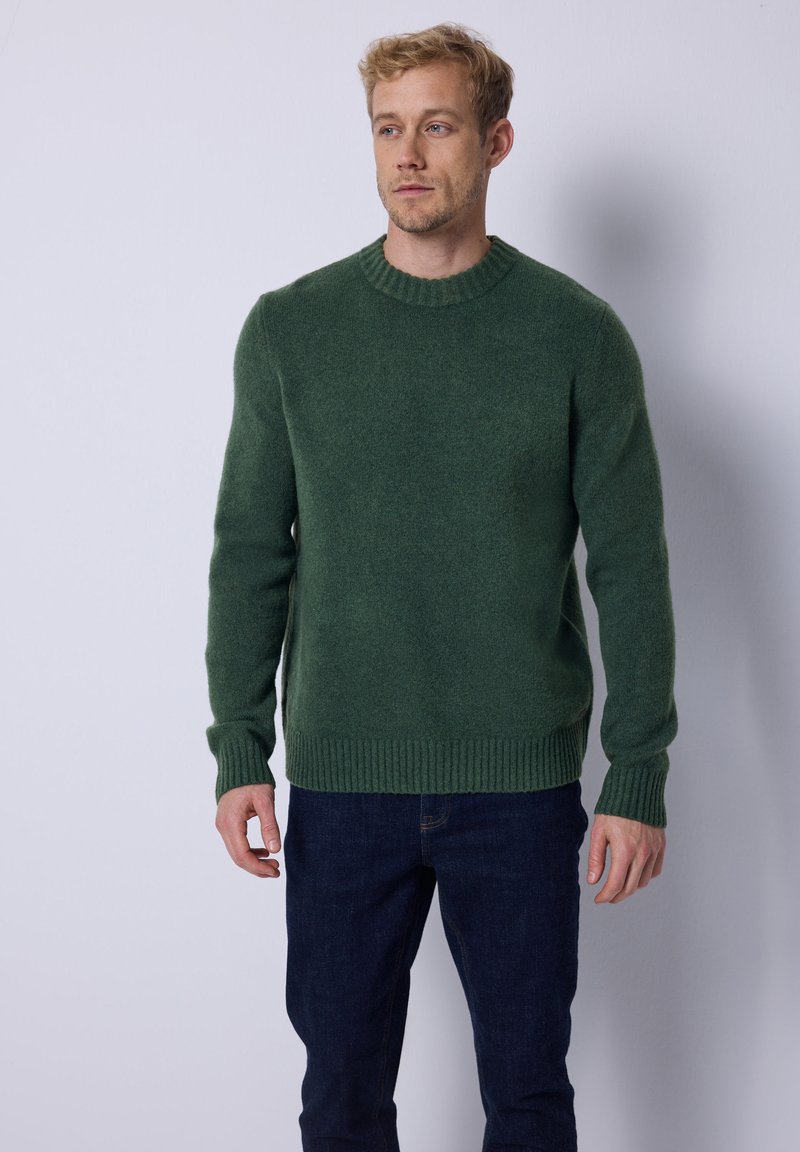 Suéter de lana verde oscuro con cuello redondo, puños y dobladillo acanalados. Combinado con jeans oscuros que presentan un diseño ajustado y una textura suave.