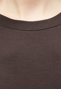 Κοντινή λήψη του λαιμού και του άνω μέρους του στήθους ενός ατόμου που φοράει ένα απλό καφέ σκούρο μπλουζάκι με στρογγυλή λαιμόκοψη.