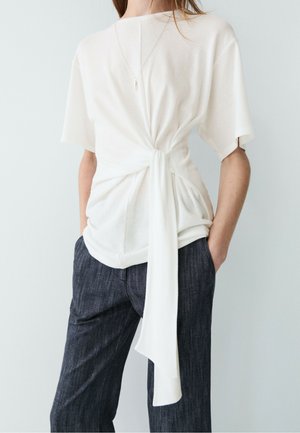 Femme portant un top blanc à manches courtes noué à la taille avec un long détail en tissu, associé à un pantalon sombre à fines rayures et un collier simple.