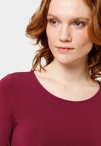 Haut bordeaux à manches longues avec un col rond, fabriqué en tissu lisse, avec un design ajusté et une texture subtile.
