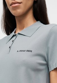 Ανοιχτό γκρι πόλο πουκάμισο με κλασικό γιακά, κούμπωμα με τρία κουμπιά και ribbed κοντά μανίκια με λεπτομέρεια μαύρου κεντημένου λογότυπου.