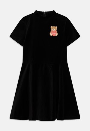 Vestido de terciopelo negro con mangas cortas, cuello redondo y falda acampanada. En el pecho, un parche de oso sosteniendo un corazón.
