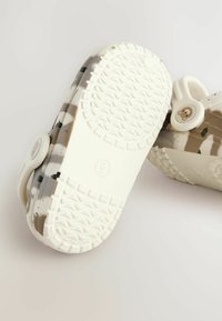 Gros plan de deux chaussures à motif camouflage, avec des semelles blanches et un logo d'ours sur la sangle, présentées sur un fond clair.