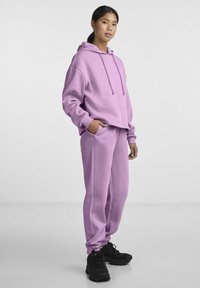 Felpa viola con cordini e vestibilità rilassata, abbinata a pantaloni jogger viola coordinati; entrambi hanno un tessuto testurizzato. Sneakers nere completano il look.