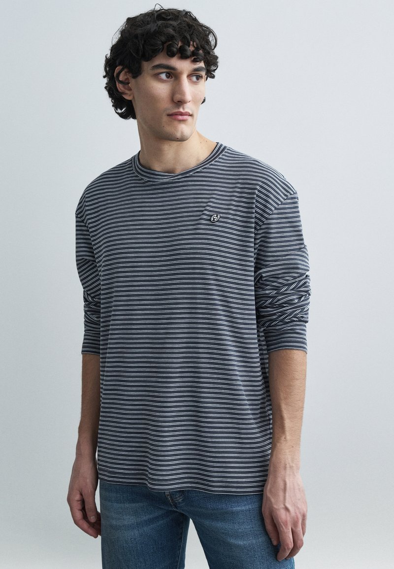 Hosszú ujjú csíkos póló sötétkék és fehér pamutból, kerek nyakkivágással, hímzett logóval a mellkason, bő szabással és felhajtott ujjakkal.