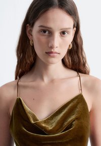 Olivengrünes Samt-Camisole mit einem drapierten Ausschnitt. Dünne Träger. Das Model trägt strukturierte goldene Creolen. Einfaches, elegantes Design.