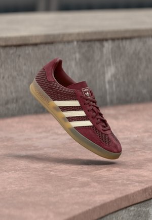 Bordowy tenisówka z materiałów siateczkowych i zamszowych. Posiada trzy białe paski, okrągły nosek i gumową podeszwę. Z tyłu logo.