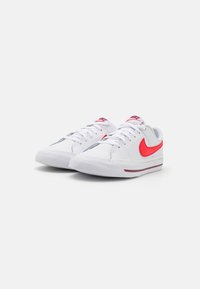 Zapatillas bajas Nike blancas con logotipos del swoosh rojos, cordones blancos y una franja color marrón en la suela, presentadas sobre un fondo blanco.