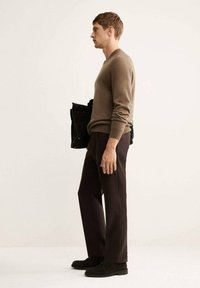 Brauner Pullover mit gerippten Bündchen, dunkelbraune Hose und schwarze Schuhe. Das Modell hält eine schwarze Tasche und steht vor einem neutralen Hintergrund.