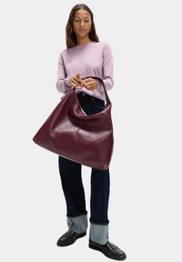 Borsa in pelle borgogna con una forma grande e morbida e un'unica maniglia curva, abbinata a un maglione viola chiaro e pantaloni di denim scuri.