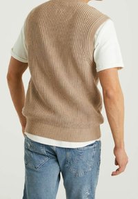 Beżowy sweter kamizelka z dzianiny, z prążkowaną teksturą i bezrękawnikowym krojem, noszony na białej koszulce, w zestawieniu z jasnoniebieskimi dżinsami.