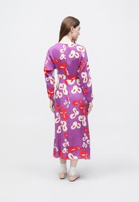 Μακρύ μωβ φόρεμα με κόκκινα και λευκά λουλουδένια σχέδια, μακριά μανίκια και φερμουάρ στην πλάτη. Το μοντέλο φοράει μπεζ σανδάλια και ανοιχτόχρωμες κάλτσες.