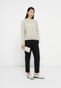 Lichtgrijze fluffy trui met "Calvin Klein" logo, zwarte op maat gemaakte broek en witte instap schoenen. Houdt een gestructureerde witte clutch vast.