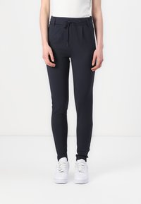Mørkeblå tapered bukser i strækbart stof med snor i taljen, forlommer og en glat tekstur; stylet med hvide sneakers.