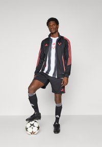 Chaqueta deportiva negra con rayas rojas, camisa blanca, pantalones cortos negros y botas de fútbol negras. De pie sobre un balón de fútbol con un patrón de estrellas.