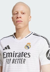 Camiseta deportiva blanca con detalles en negro y texto "Emirates Fly Better". Presenta un cuello acanalado y un escudo del club con una corona en el lado izquierdo del pecho.