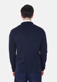 Maglione a maniche lunghe color navy con texture a coste, colletto a scialle e taglio dritto, indossato sopra una camicia con colletto e abbinato a jeans in denim.