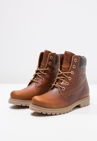 Botines de cuero marrón con punta redonda, suela de goma beige y cierre de cordones en la parte delantera. Presentan detalle de costuras y logotipo en el lateral.