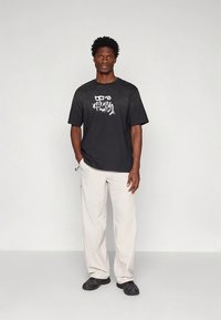 Zwart T-shirt met korte mouwen en een grafisch ontwerp aan de voorkant, gecombineerd met lichtbeige losse broek en zwarte schoenen.