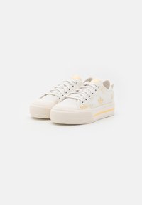 Vita canvas sneakers med ljusgula detaljer, rund tå, texturerad gummisula och ett subtilt blommönster på sidan.