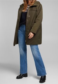 Chaqueta parka verde oliva con capucha, bolsillos laterales y cierre frontal con cremallera. Combinada con jeans acampanados de color azul claro y botines negros.