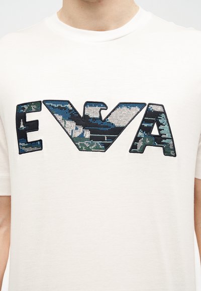 Λευκό μπλουζάκι με μεγάλα κεντημένα γράμματα "EVA" σε μοτίβο καμουφλάζ με μπλε, πράσινο, μαύρο και λευκό χρώμα στην περιοχή του στήθους.