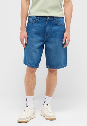 Mann trägt blaue Jeans-Shorts mittlerer Länge, weiße Socken mit schwarzem Text und weiße Freizeitsneakers, steht vor einem einfarbigen Hintergrund.