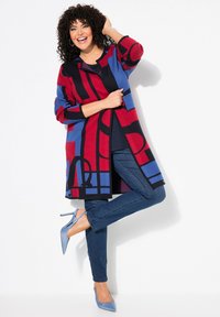 Cardigan en tricot avec des motifs géométriques rouges, bleus et noirs ; associé à un jean foncé et des talons aiguilles bleu clair. Manches longues et ouverture devant.