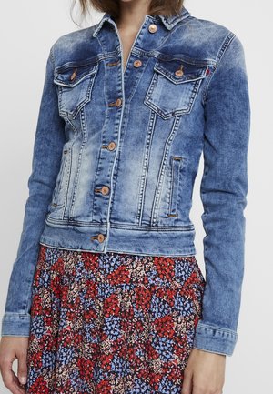 Veste en denim délavé bleue avec deux poches sur la poitrine, des boutons en métal et un design ajusté. Portée sur une jupe à motifs floraux.