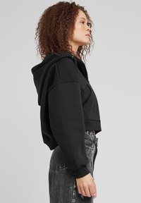 Felpa nera corta con cappuccio, dotata di maniche lunghe, polsini a coste e design minimalista. Indossata con jeans a vita alta e sbiaditi.