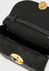 Schwarze Lederhandtasche mit Wildlederklappe, rundem goldfarbenem Verschluss und einem Reißverschlussfach im Inneren, offen dargestellt vor weißem Hintergrund.