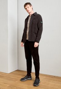 Grey corduroy jacket with front pockets, beige T-shirt, black corduroy pants, and grey slip-on shoes with textured soles. Wooden floor background.