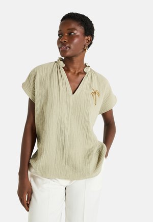 Blusa de textura verde claro con escote en V, cuello con volantes arrugados y detalle de bordado de palmera. Llevada con pantalones blancos.