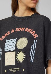 Schwarzes, übergroßes T-Shirt mit grafischem Druck, das Sonnenmotive und Wellen zeigt. Der Text enthält "NIMM DIR EINE SONNENPAUSE" und "EST 94" in verschiedenen Farben.