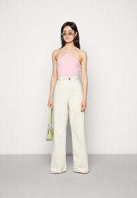 Top halter rosa e calças largas em tom creme. Modelo segura uma mala texturizada verde. Óculos de sol acrescentam um detalhe moderno. Fundo simples.