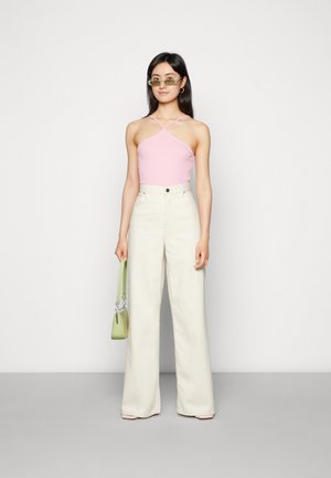 Top halter rosa e calças largas em tom creme. Modelo segura uma mala texturizada verde. Óculos de sol acrescentam um detalhe moderno. Fundo simples.