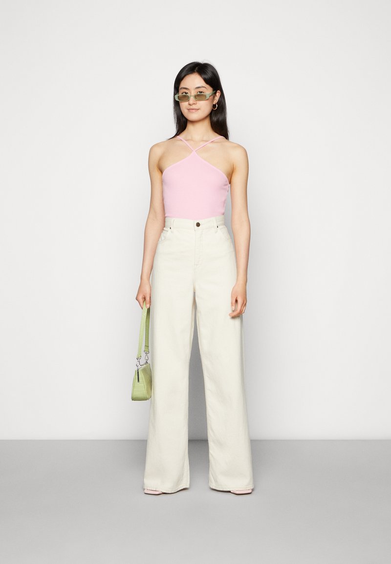 Top halter rosa e calças largas em tom creme. Modelo segura uma mala texturizada verde. Óculos de sol acrescentam um detalhe moderno. Fundo simples.