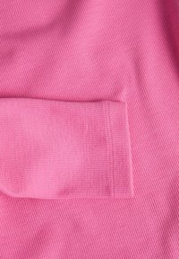 Růžová jemně vroubkovaná tkanina s texturovaným povrchem, obsahující detail kapsy na levé straně a hladké zakončení podél okrajů.