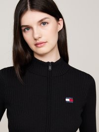 Top nero a coste con zip e colletto alto, caratterizzato da una piccola patch del logo "Tommy Jeans" in rosso, bianco e blu sul lato sinistro.