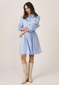 Mujer con vestido azul claro con detalles de encaje en las mangas y el pecho, combinado con botas beige hasta la rodilla, posando contra un fondo liso.