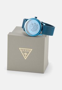 Reloj con correa de malla azul y caja redonda, cara texturizada de color azul claro con un diseño de león, exhibido sobre una caja gris con logo dorado.